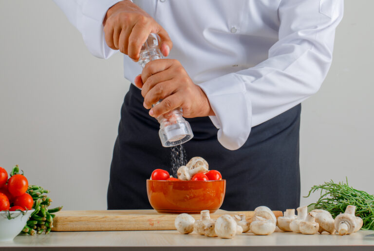 male chef adding salt into food in kitchen in uniform and apron front view.